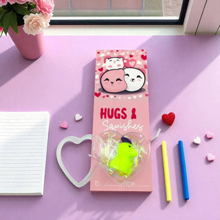 Pink packaging of 'Hugs & Squishies' with a green toy inside, on a pink surface with heart decorations.