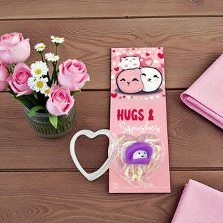 Pink packaging of 'Hugs & Squishies' with a heart-shaped cookie cutter and flowers on a wooden surface