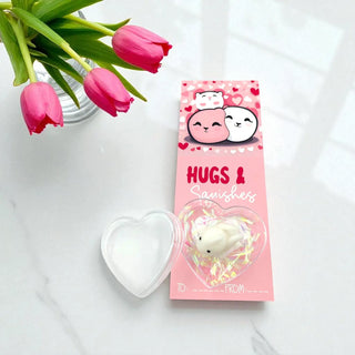 Squishy toy in a heart-shaped packaging with 'Hugs & Squishies' text, next to pink tulips on a white surface.