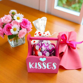 Pink 'Kisses' container with candy, flowers, and a pink bow on a wooden surface.