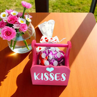 Pink 'Kisses' basket with wrapped candies and a vase of flowers on a wooden table.