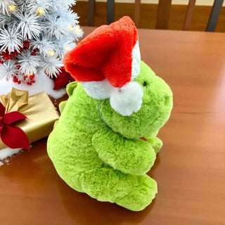 Green plush toy with a red Santa hat on a wooden table with Christmas decorations.