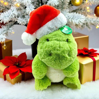 Green plush dinosaur toy with a Santa hat in front of a Christmas tree and presents.