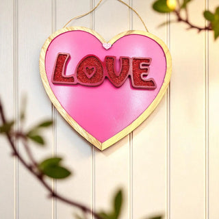 Heart-shaped sign with 'LOVE' in red on a pink background, hanging against a wooden wall.