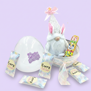 Easter-themed gift set with a plush bunny, egg-shaped container, and candy on a light purple background.