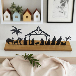 Silhouette nativity scene on a wooden board with decorative houses and plants in the background.
