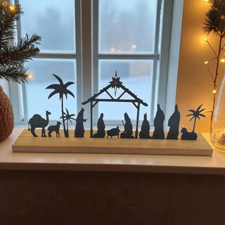Silhouette nativity scene on a wooden stand with Christmas decorations in the background.