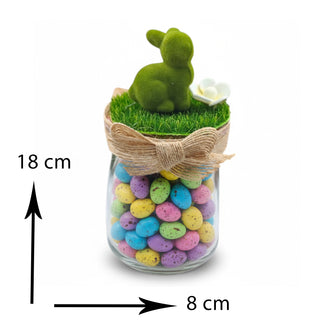 Decorative jar with pastel-colored stones, grass, and a green figurine on a white background.