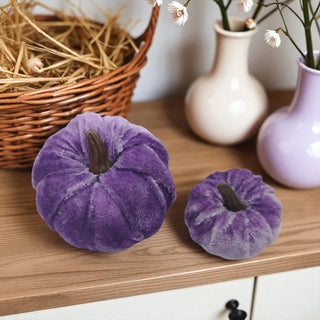 Two purple velvet pumpkins on a wooden surface with decorative vases and hay in the background.