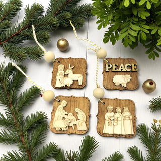 Wooden Christmas ornaments with nativity scene and 'PEACE' design, surrounded by pine branches and gold balls.