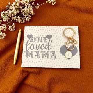 Card with 'One Loved Mama' text, pen, and keychain on an orange fabric background