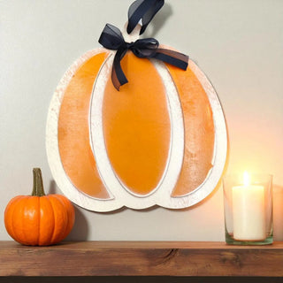 Decorative pumpkin wall art with a black ribbon on a wooden shelf with a small pumpkin and candle.