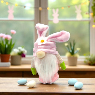 Decorative gnome with bunny ears on a table with Easter eggs and flowers in the background.