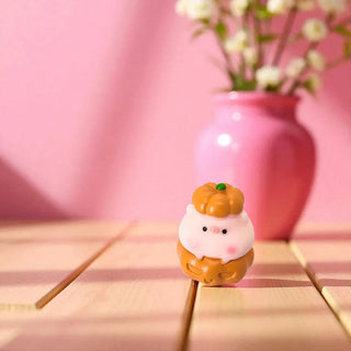 Small decorative item resembling a cartoon character with a pumpkin on a wooden surface with a pink background.