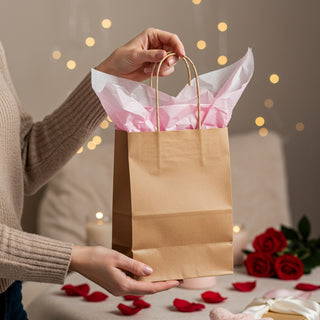 Person holding a brown paper bag with pink tissue paper against a warm, blurred background.