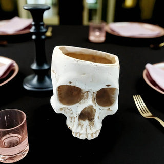 Skull-shaped ceramic mug on a table with a black tablecloth and pink glasses.