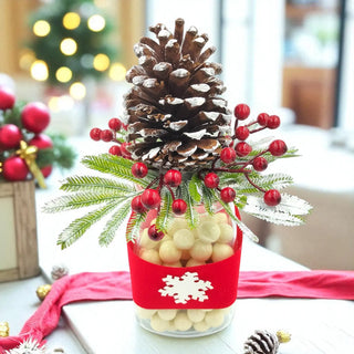 Decorative Christmas arrangement with pine cones, berries, and a red bag on a table.