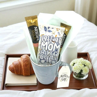 Tray with coffee, croissant, and flowers on a bed, featuring a 'Mothers Day' tag.