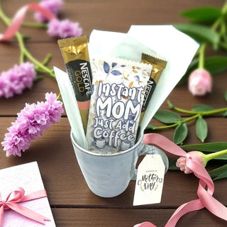 Mug with coffee packets labeled 'Instant Mom' and 'Nescafe Gold', surrounded by flowers and a gift box on a wooden surface.