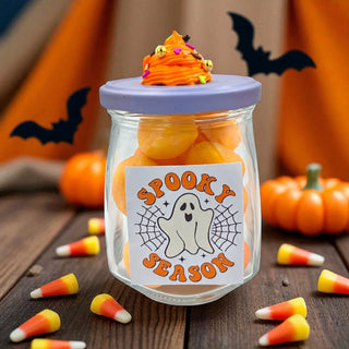 Glass jar with Halloween-themed candy and decorative label on a wooden surface with pumpkins and bats in the background.