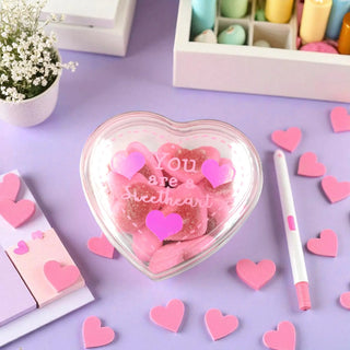 Heart-shaped container with pink candies and heart decorations on a purple surface