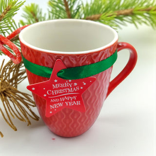 Red mug with a green ribbon and 'Merry Christmas and Happy New Year' tag, surrounded by Christmas decorations.