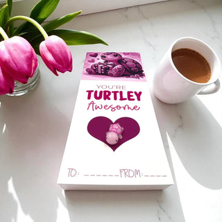 Card with 'You're Turtley Awesome' message on a marble surface with a cup of coffee and pink tulips.