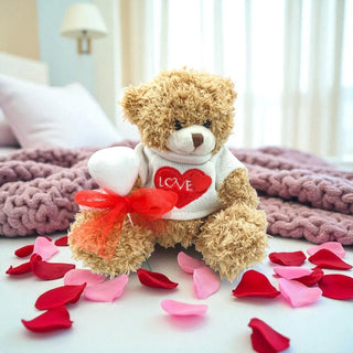 Teddy bear wearing a 'LOVE' shirt with red and pink ribbons on a bed with rose petals.