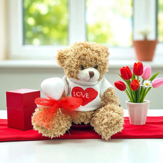Teddy bear with a heart-shaped 'LOVE' shirt on a red tablecloth with flowers and a gift box.