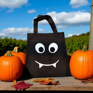 Black tote bag with a smiley face design surrounded by pumpkins and leaves outdoors.
