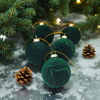 Green velvet Christmas ornaments with gold ribbons on a snowy surface with pine branches and cones.