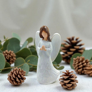 Angel figurine with brown hair surrounded by pine cones and greenery on a light background