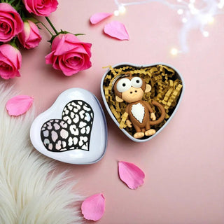 Heart-shaped box with a monkey figurine, surrounded by pink roses and petals on a pink background.