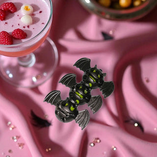 Three bat-shaped pins on a pink surface with a cocktail in the background