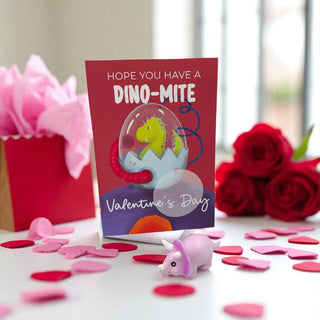 Valentine's Day card with dinosaur design, small pink toy, and flowers on a table.