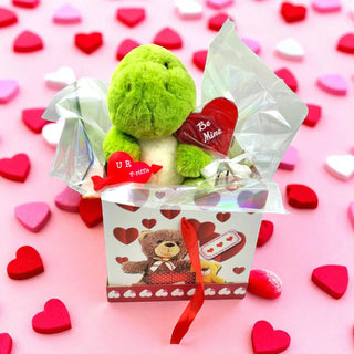 Green plush toy with heart-shaped tag in a gift box on a pink background with heart decorations
