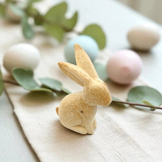 Small rabbit figurine on a wooden surface with pastel Easter eggs and greenery.