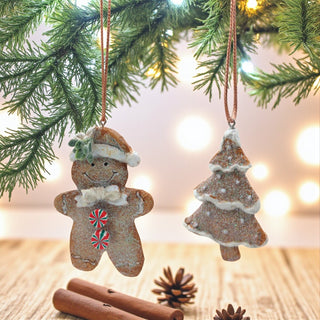 Two Christmas ornaments shaped like a gingerbread man and a tree hanging from a tree branch with lights in the background.