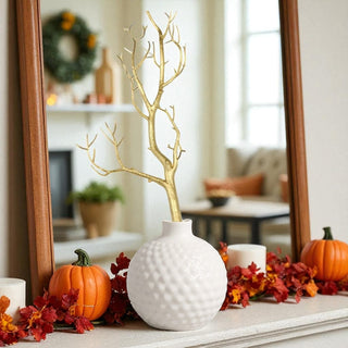 Decorative setup with a white textured vase, gold branch, pumpkins, and fall leaves in a home setting.