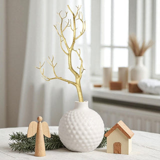 Decorative setup with a white vase, gold branches, wooden angel, and house on a light surface.