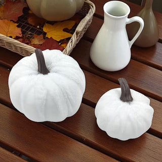 Two white pumpkins on a wooden surface with a white vase and leaves in the background.
