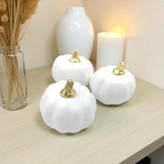 Three white pumpkins with gold stems on a table next to a vase with dried plants and a candle.