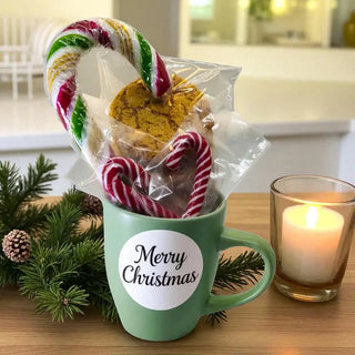 Green mug with 'Merry Christmas' label, candy canes, cookies, and a candle on a wooden surface.