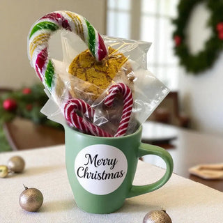 Green mug with 'Merry Christmas' label filled with cookies and candy canes on a festive table.