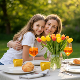 Woman and young girl sitting at a table with tulips and drinks, smiling outdoors.