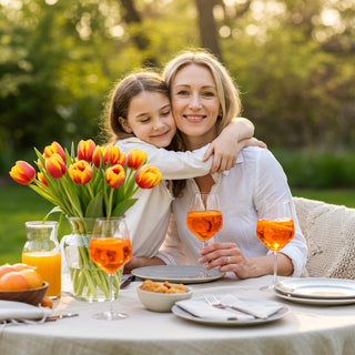 Woman and young girl hugging at a table set with orange juice, snacks, and tulips outdoors.