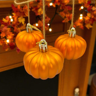 Three small orange pumpkins hanging against a festive background with lights and autumn leaves.