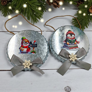 Two Christmas-themed bottle cap ornaments with penguins and snowflakes on a wooden surface with festive decorations.