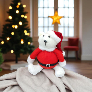 White teddy bear in a Santa outfit on a blanket with a Christmas tree in the background