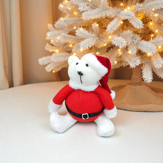 White teddy bear in a Santa outfit sitting in front of a decorated Christmas tree.